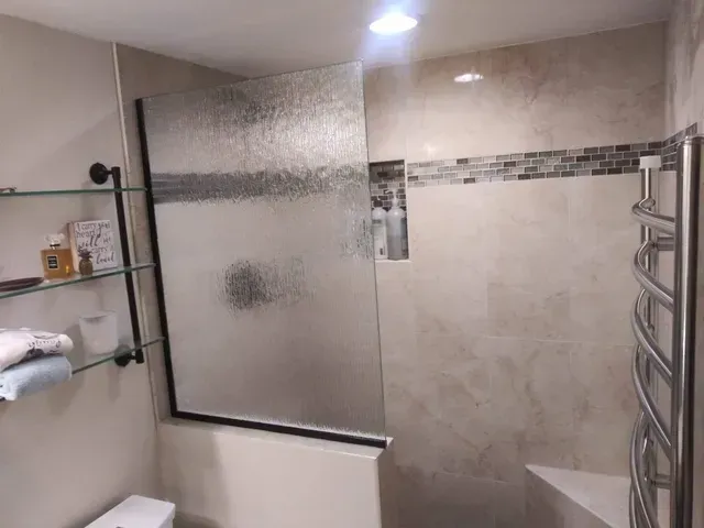 A bathroom with a textured glass shower panel, a glass shelf unit, and a chrome towel warmer mounted on a tiled wall.