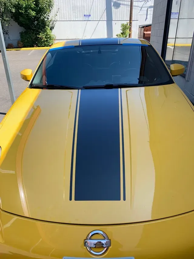 Yellow Nissan car hood with a black racing stripe, shown from a front-facing perspective outdoors.