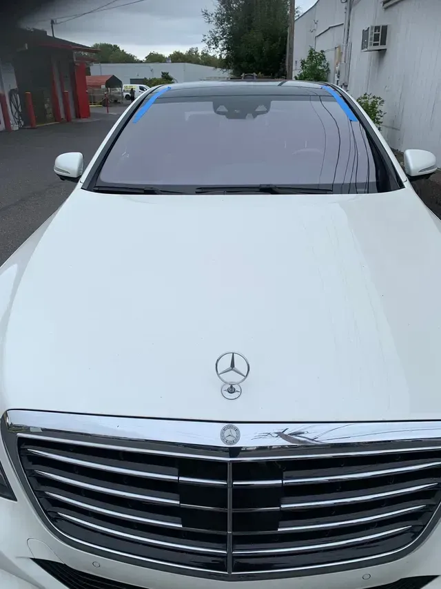 A front-facing view of a white Mercedes-Benz sedan parked on pavement with blue masking tape along the windshield edge.