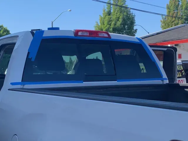 The rear window of a white pickup truck is outlined with blue painter's tape, likely in preparation for window tinting.