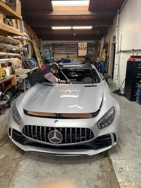 A mechanic working on the windshield of a silver Mercedes-Benz sports car inside a cluttered auto repair shop.