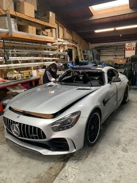 A mechanic works on a silver Mercedes-AMG GT sports car inside an automotive repair shop with storage shelving.