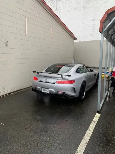 Silver Mercedes-AMG GT sports car with a rear spoiler parked in a paved lot next to a white brick building.