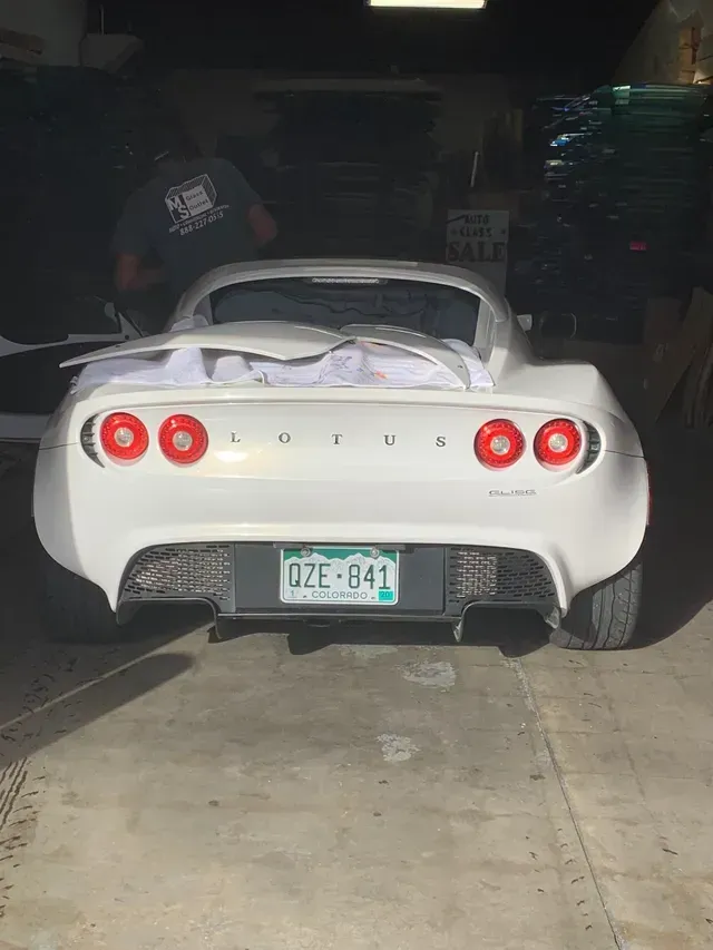 A white Lotus sports car is parked inside a dim garage, viewed from the rear with a license plate visible.