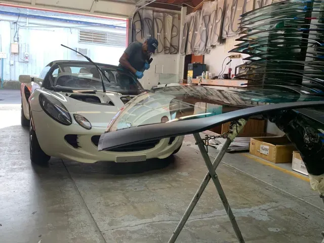 A technician works on a white Lotus sports car inside a shop with a new windshield prepared on a stand.