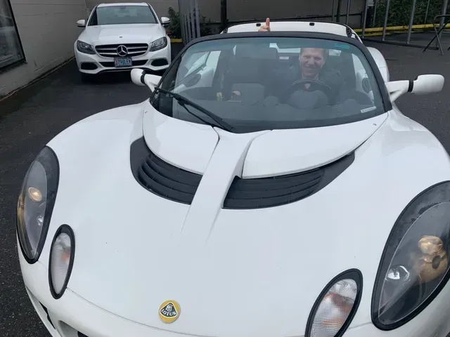 A smiling person in a white Lotus convertible giving a thumbs-up, with a white Mercedes parked in the background.