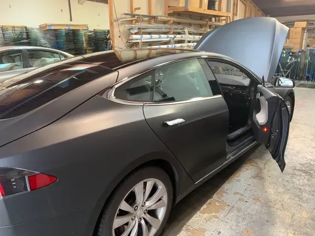 A matte charcoal Tesla Model S parked inside a workshop with its hood and front passenger door open.