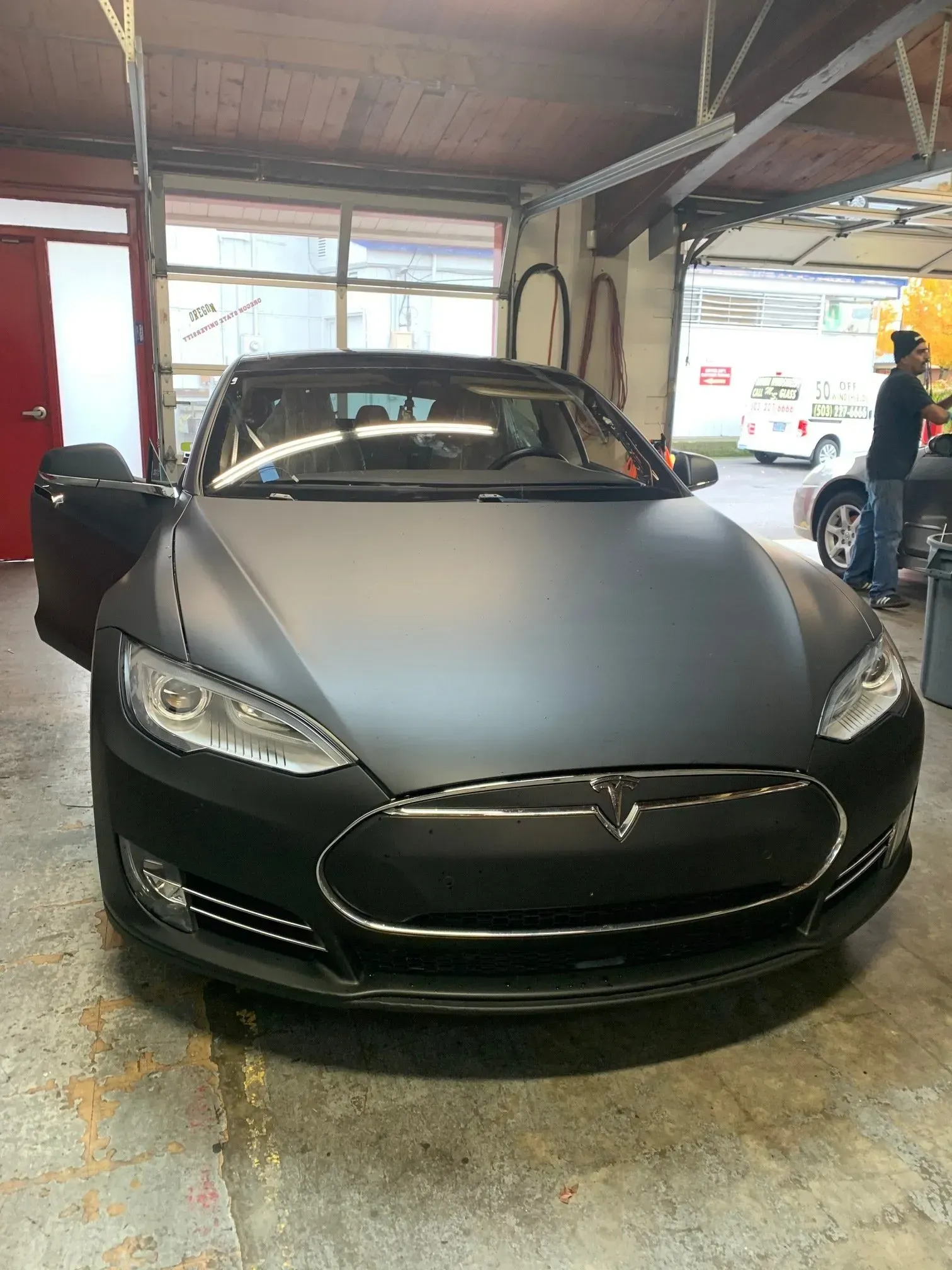 A matte black Tesla Model S parked inside a workshop with a person standing by a car in the background.