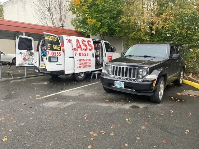 A white service van with open doors and a black SUV parked in an outdoor parking lot with trees in the background.