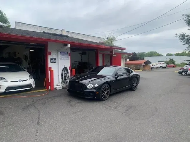 A black luxury sports car parked in front of a service station, with a white car partially visible in the garage bay.