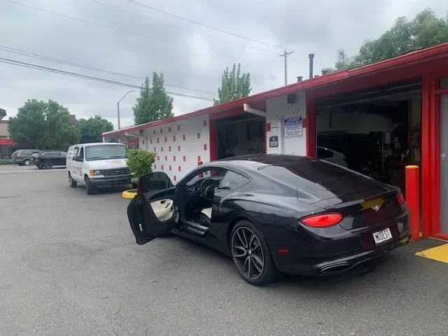 A black Bentley with an open door parked in front of a building with red trim, with a white van in the background.