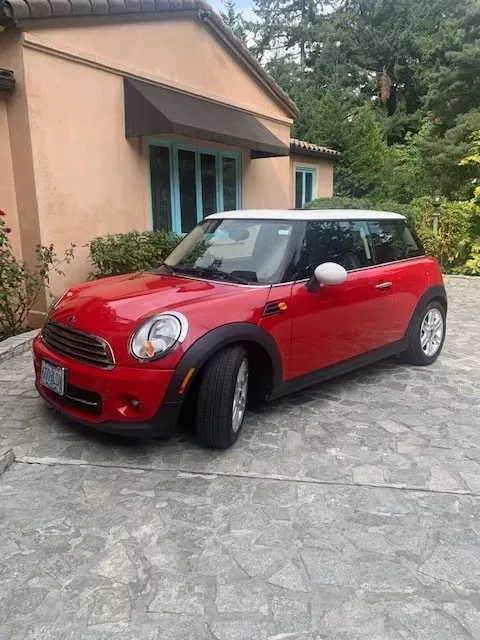 A red Mini Cooper with a white roof parked on a stone driveway in front of a house.