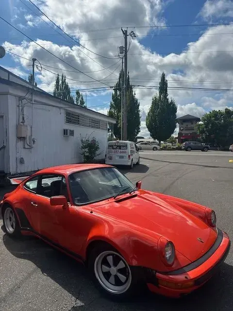 A bright orange Porsche 911 parked in an outdoor lot on a sunny day with a white building and trees in the background.