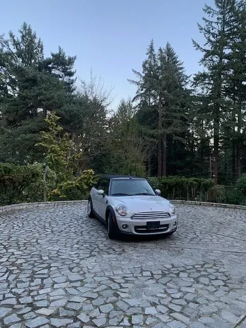 A white Mini Cooper convertible parked on a stone-paved driveway surrounded by tall trees at dusk.