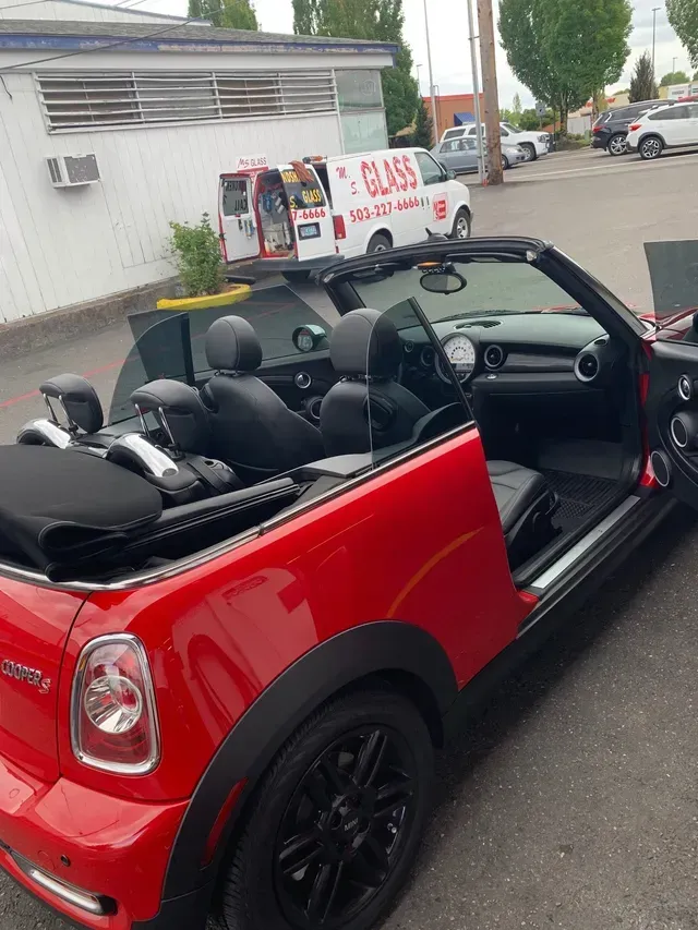 A red Mini Cooper convertible parked outdoors, with a glass repair van in the background.