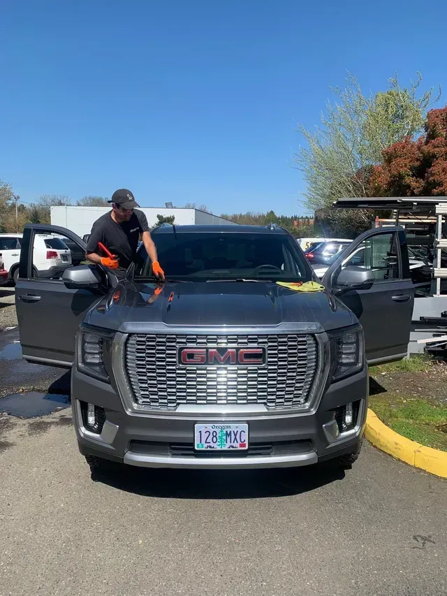 A technician wearing orange gloves works on the windshield of a dark grey GMC SUV parked in an outdoor lot.