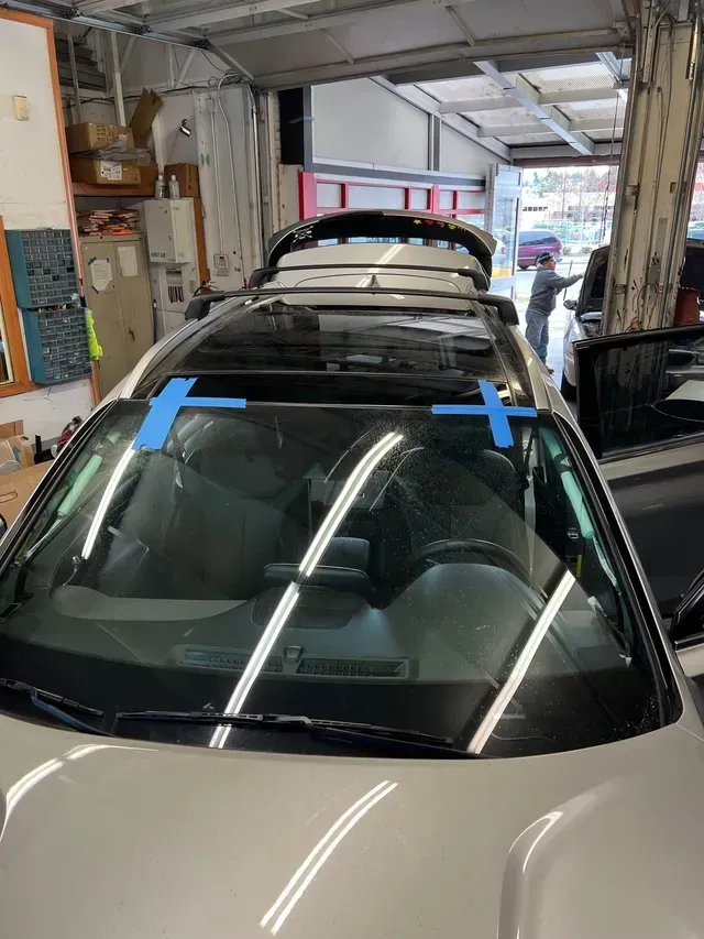 A light-colored car in a garage with blue painter's tape on the windshield and a person working in the background.