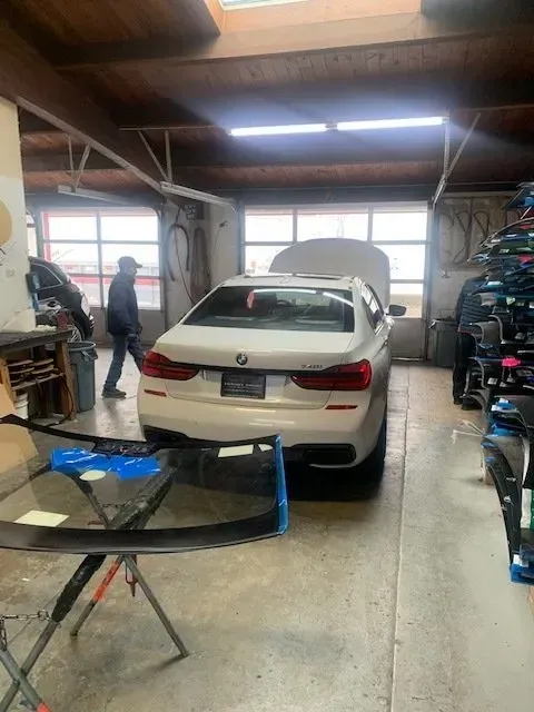 A white BMW sedan with its hood open in a garage, with workers nearby and a car windshield on a stand in the foreground.