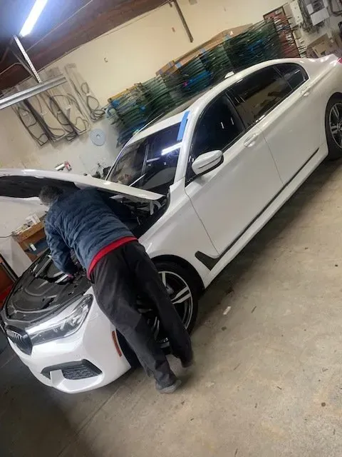 A person in a blue jacket works on the engine of a white sedan inside a garage.
