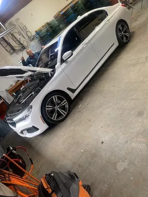 A white BMW sedan with its hood open in a workshop, with a person standing behind the open driver-side door.