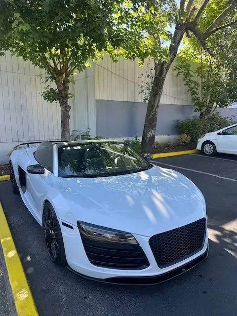 A white Audi R8 convertible parked in a paved lot under trees.