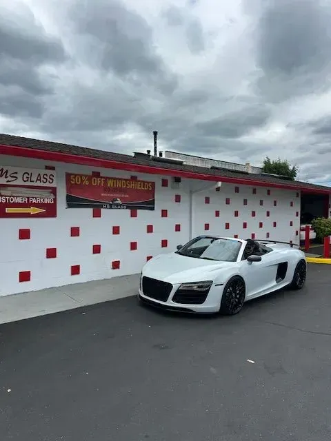A white Audi R8 convertible parked in front of a building with a white facade featuring red square accents.