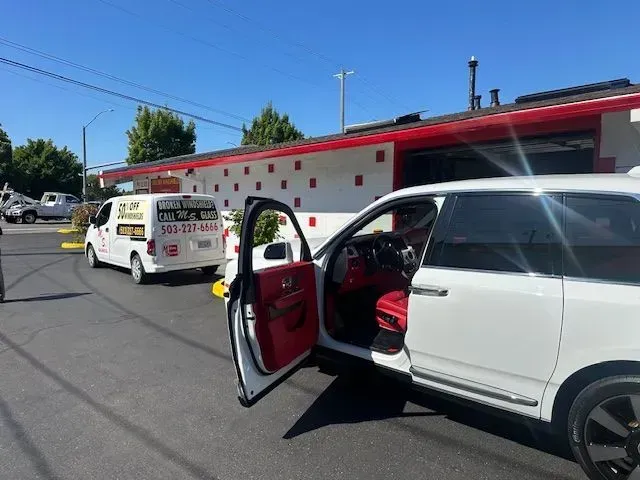 A white minivan with open doors is parked in front of a white building with red trim, next to a service van.