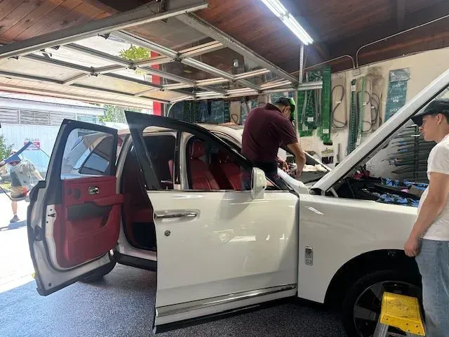 Mechanics work on a white SUV with open doors and a raised hood in a garage.
