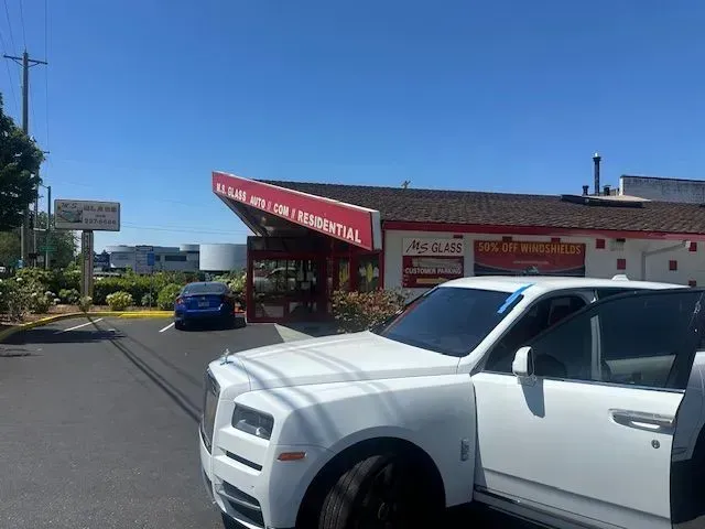 A white Rolls-Royce with an open door parked in front of a glass repair business under a clear blue sky.