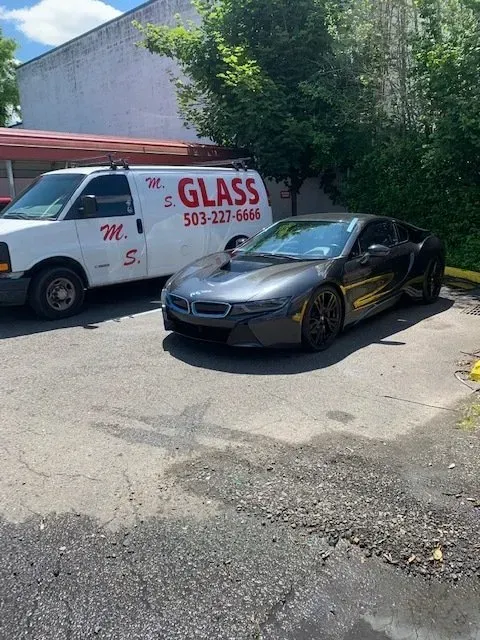 A dark gray BMW i8 parked next to a white M.S. Glass service van in an outdoor lot on a sunny day.