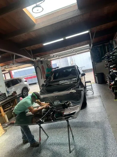 A worker in a light green shirt repairs a car hood on a stand inside a well-lit auto shop, with another car visible nearby.