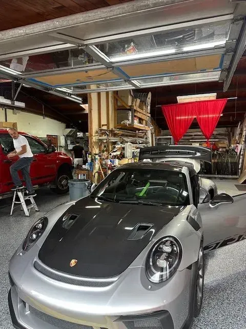 A person stands on a ladder working on a red truck in a shop next to a silver and black Porsche.
