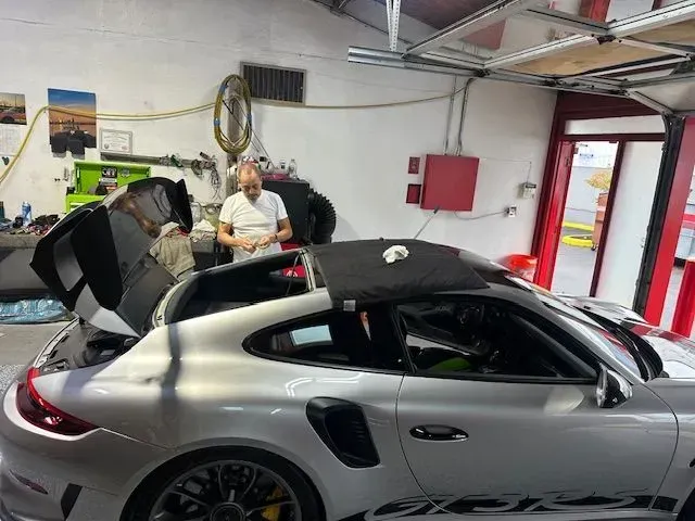 A silver Porsche sports car with its rear engine cover open, being worked on by a person inside an auto repair garage.
