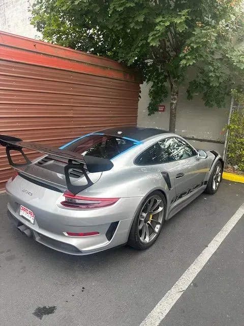 A silver Porsche 911 GT3 RS with a prominent rear spoiler, parked in a lot next to a corrugated metal wall.