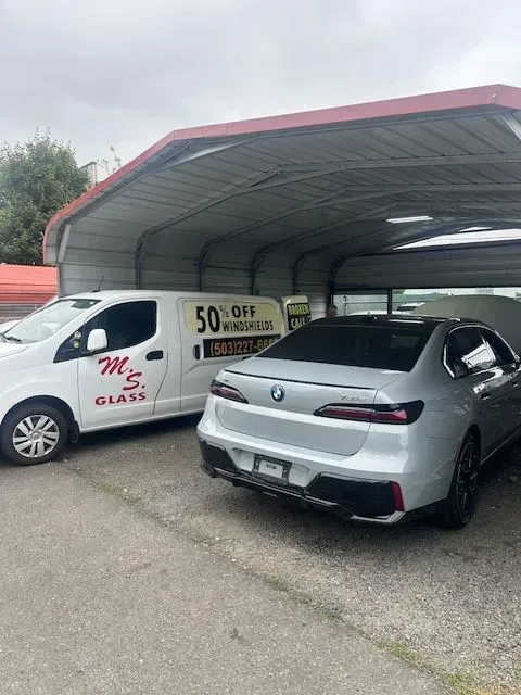 A silver BMW sedan parked next to a white M.S. Glass service van under a metal carport.