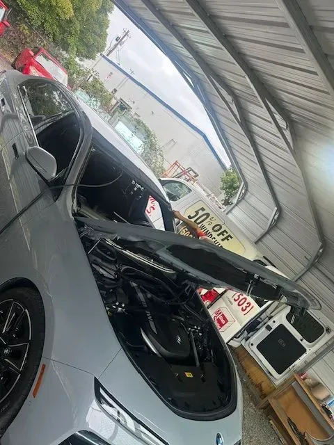 A light gray car parked under a metal canopy with its hood raised, showing the engine compartment.