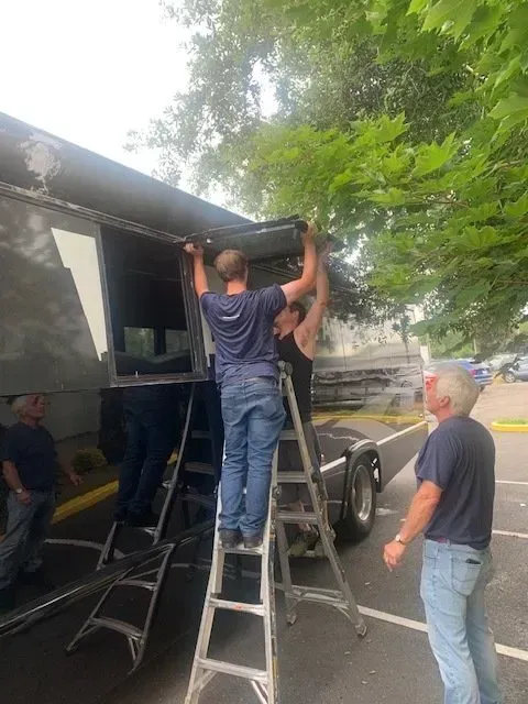 Four people work together to install or repair a large metal panel on the side of a parked bus outdoors.