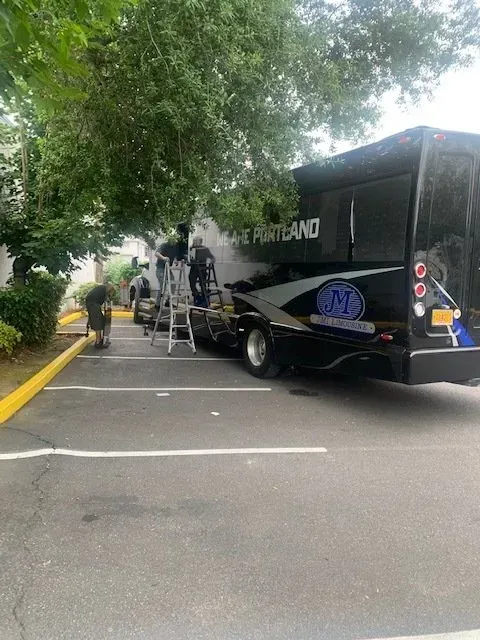 Two people on ladders perform maintenance on the side of a black coach bus parked in an outdoor lot.