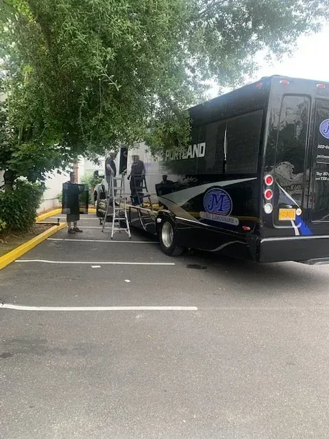 Three people stand near a black specialty vehicle in a parking lot, one using a ladder to work on a building exterior.