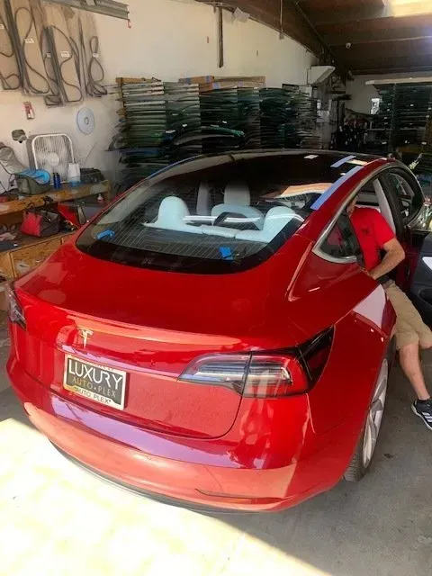 A red Tesla parked inside a shop with stacks of glass in the background; a person is sitting in the driver's seat.