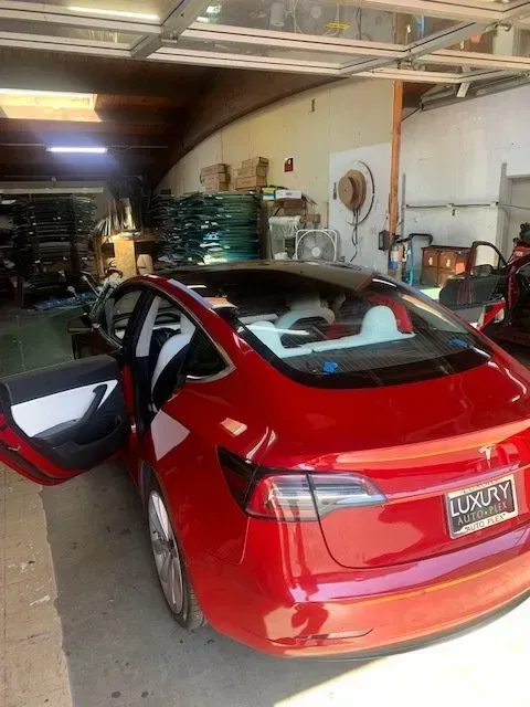 Red Tesla Model 3 parked in a garage with its driver-side door open and white interior visible.