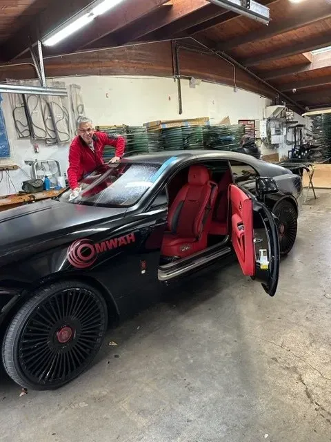 A person in a red jacket leans over a black coupe with red interior, parked inside a garage.