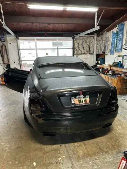 Black sedan with its driver-side door open, parked inside a cluttered garage under bright overhead lighting.