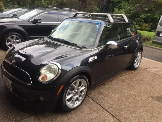 A dark-colored Mini Cooper with a silver roof rack parked on a concrete driveway.