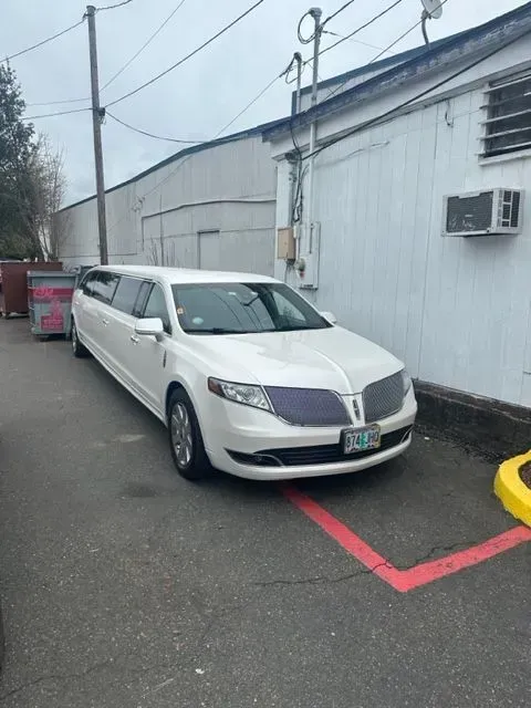 A long, white Lincoln stretch limousine is parked in an asphalt lot next to a white building.