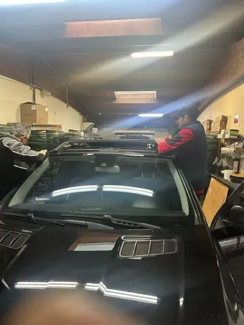 Two people work on the roof of a black vehicle inside a cluttered garage workshop.