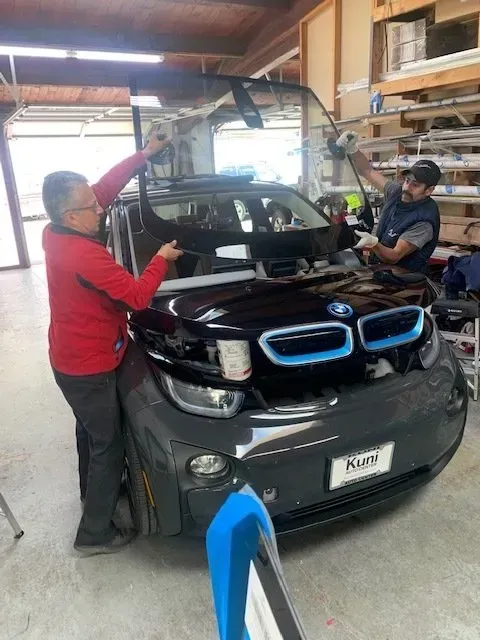 Two technicians work in a garage to install a new windshield on a dark-colored BMW i3 electric car.