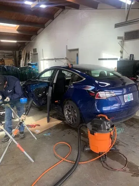 A mechanic works on the door panel of a dark blue Tesla parked in a garage, next to a large orange shop vacuum.