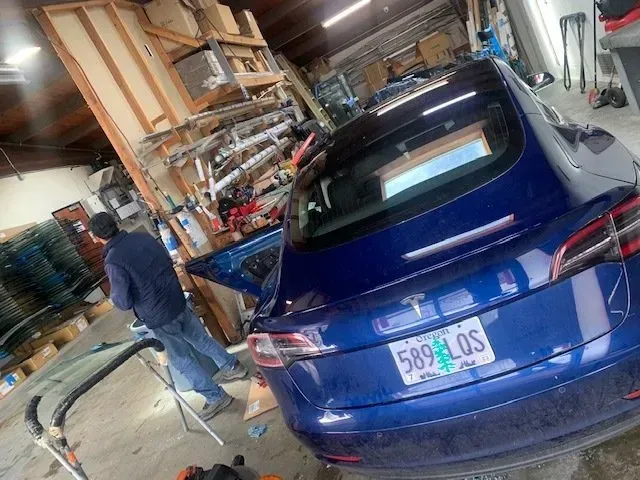 A blue Tesla parked in a cluttered workshop while a person in a dark jacket stands beside the open driver-side door.