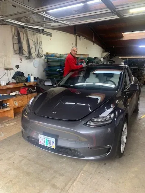 A person in a red jacket stands inside a garage, detailing the windshield of a dark grey Tesla vehicle.